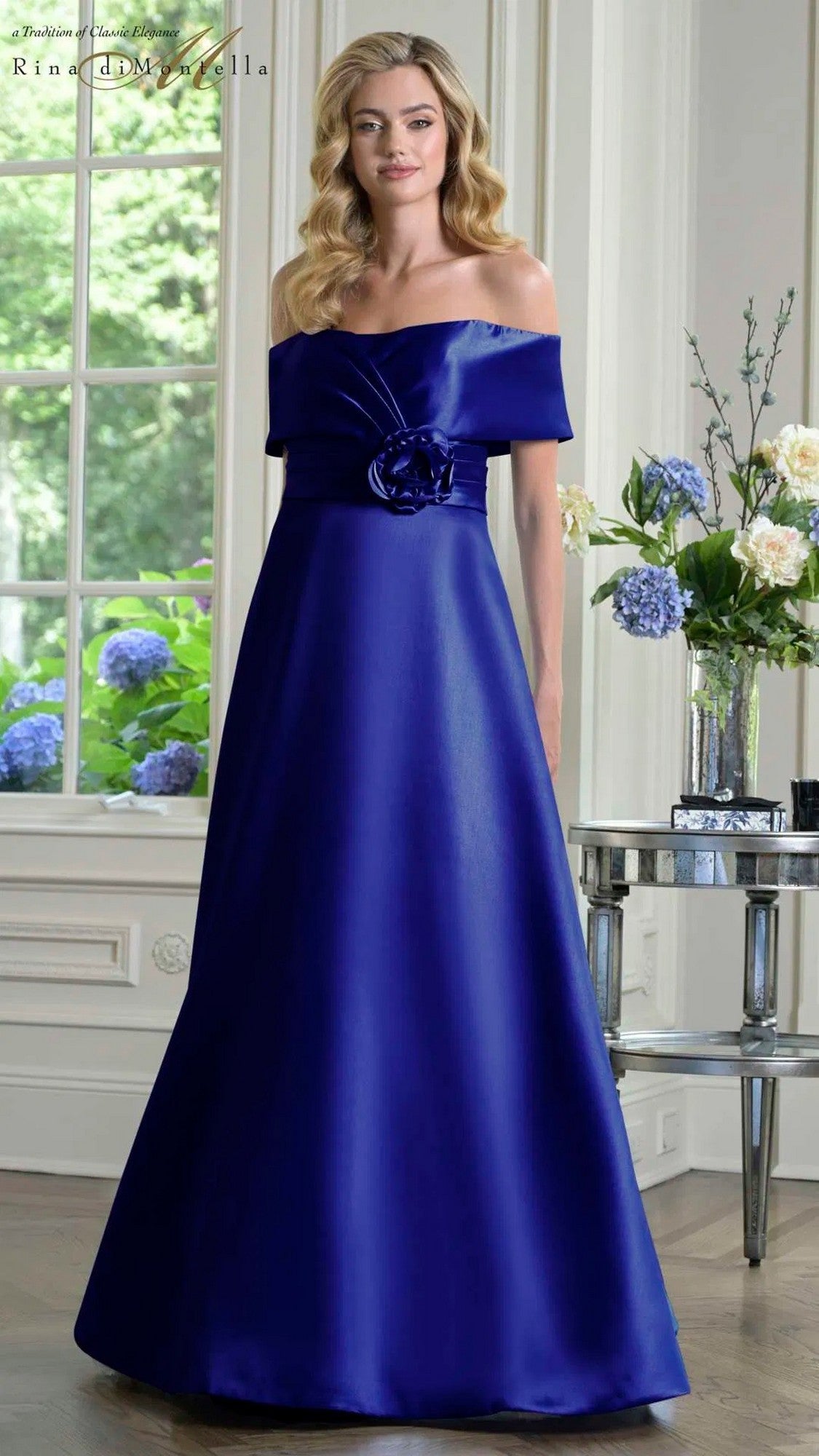 A woman stands indoors in the Rina Di Montella RD3429 long formal dress, featuring navy Mikado fabric with an off-the-shoulder neckline and a floral waist accent.